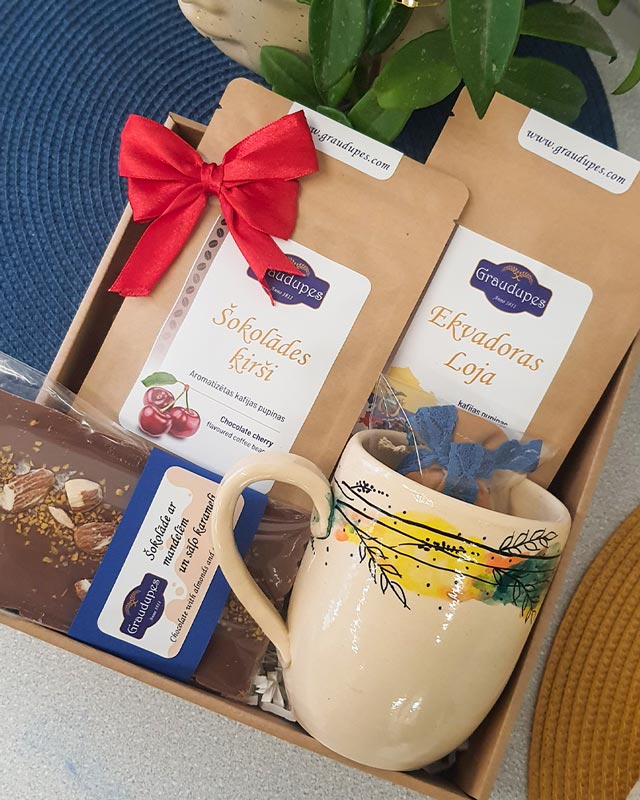 The "Coffee Mug" gift set includes two 100g packets of 100% Arabica coffee, one Cherry Chocolate flavored and one Specialty Ecuador, a milk chocolate bar with almonds and salted caramel, and a unique hand-painted mug, all in an elegant gift box.