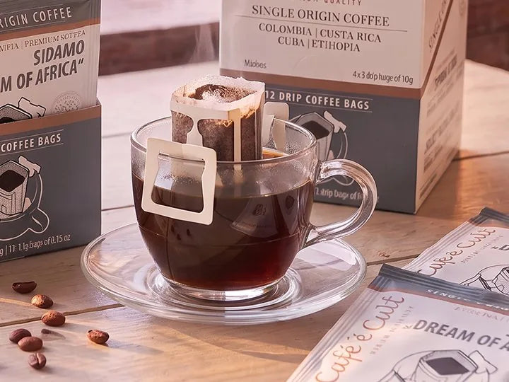 A drip coffee bag hanging on a glass cup of fresh black coffee, with coffee beans and drip sachets on a wooden table